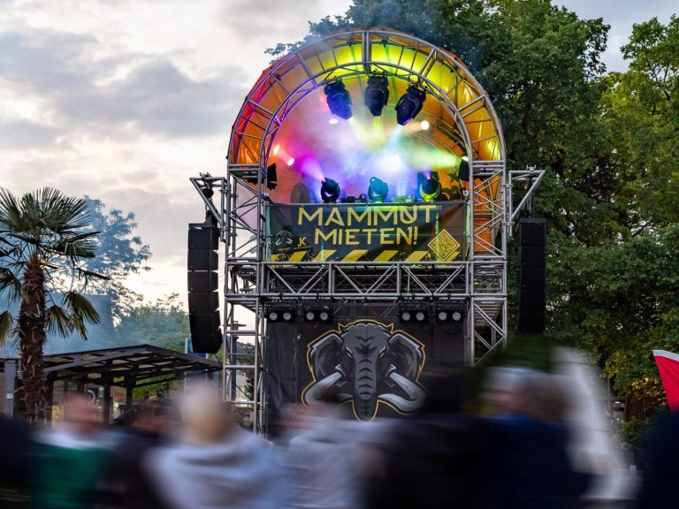 Bunte Lichter blitzen von einer großen Lautsprecheranlage auf einem Festivalgelände.