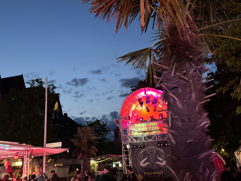 Bunte Neonlichter unter einem Abendhimmel mit Palmen und Menschen auf einer Veranstaltung.